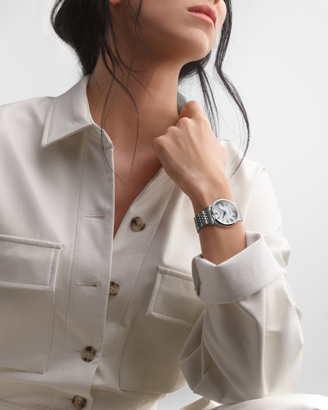 A close-up of a woman holding his hand to her jacket collar with a watch around his wrist. The watch features a silver dial, black & blue hands and markers, a stainless steel bezel, a crown on the right side, and a brown leather strap.