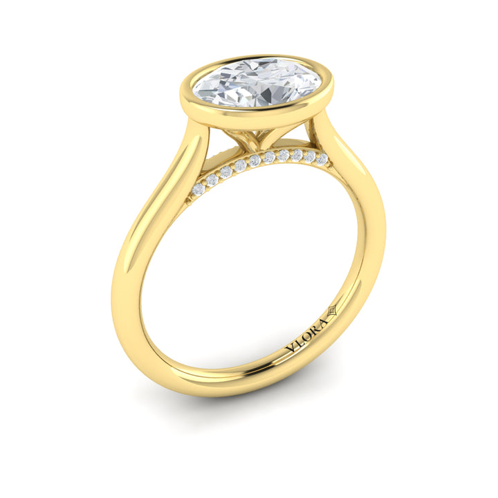 An angled view of a yellow gold engagement ring is set against a white background. The engagement ring features a bezel-set east-west oval diamond center stone, cradled in a cathedral arch, and has subtle pavé detailing around the yellow gold band.
