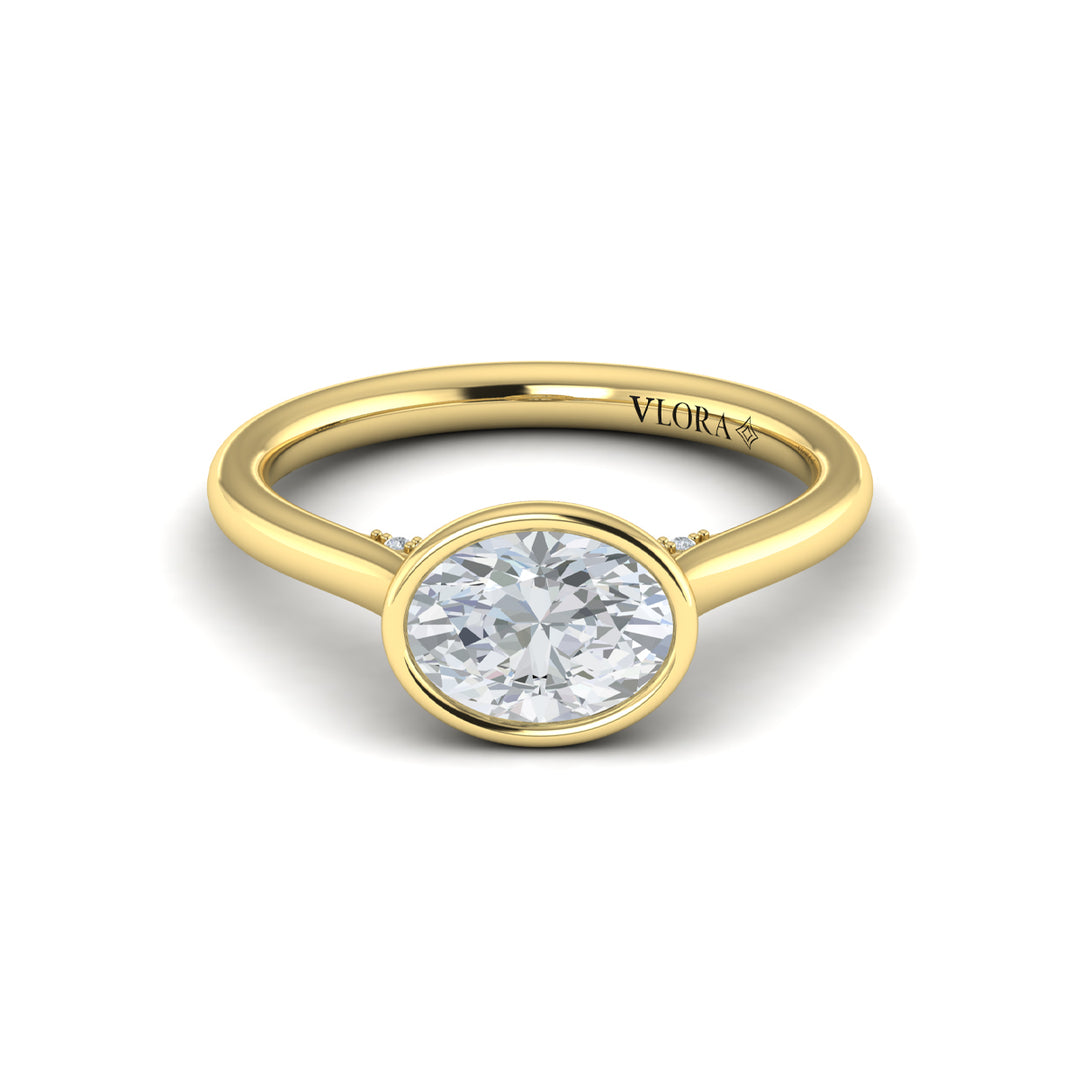 A front view of a yellow gold engagement ring is set against a white background. The engagement ring features a bezel-set east-west oval diamond center stone, cradled in a cathedral arch, and has subtle pavé detailing around the yellow gold band.