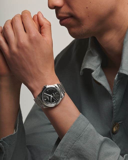 LONGINES Conquest watch with a dark dial and stainless steel bracelet worn by a man in a dark green shirt.