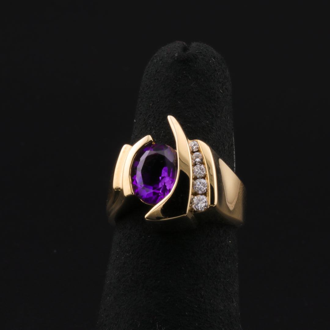 A yellow gold ring displayed in the middle of a black background. The ring features an oval-shaped purple center stone and five round-shaped side diamonds.