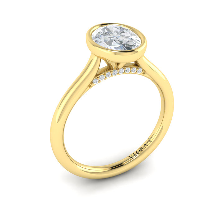 An angled view of a yellow gold engagement ring is set against a white background. The engagement ring features a bezel-set oval diamond center stone with a cathedral setting, and has subtle pavé detailing around the yellow gold band.