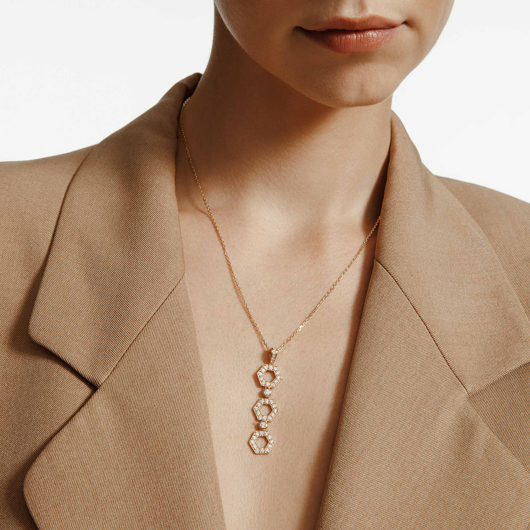 A close-up of a woman wearing a khaki blazer and a yellow gold diamond pendant necklace. The ring features a drop design with shaped hexagons connected by round bezel-set diamonds.