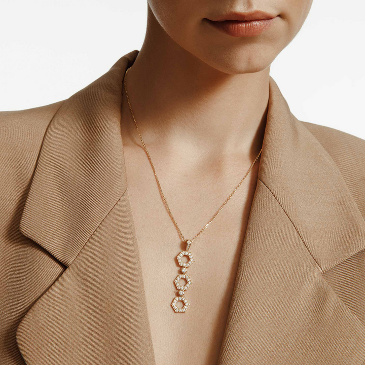 A close-up of a woman wearing a khaki blazer and a yellow gold diamond pendant necklace. The ring features a drop design with shaped hexagons connected by round bezel-set diamonds.