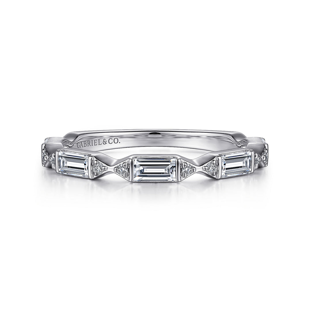 A white gold ring rests in the middle of a white background. The ring features a half-eternity style of emerald-shaped stations, prong-set round-shaped side stones, and Gabriel & Co engraved inside the ring on the left side.