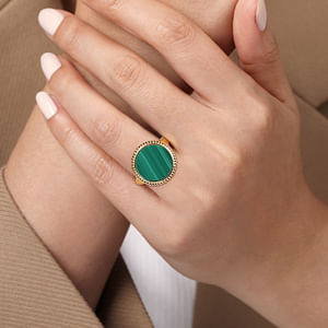 A close-up of a woman's hand showcasing the yellow gold ring on her ring finger. The ring features an inlay of vibrant green malachite gemstone and yellow gold Bujukan beading.