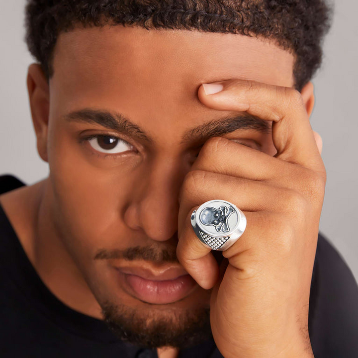 A close-up of a man holding his hand over his face, showcasing the sterling silver ring on his pinky finger. The ring features a skull motif that forms this Sterling silver signet ring.