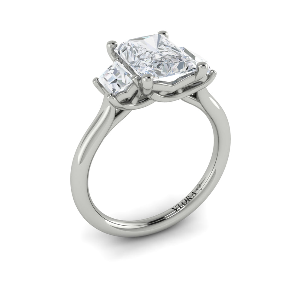 An angled view of a white gold engagement ring is set against a white background. The engagement ring features an emerald cut center diamond, flanked by complementary emerald cut side stones, in a woven setting. 