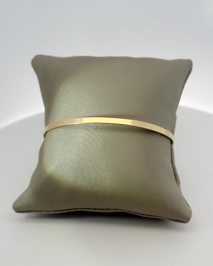 The Essential 14K yellow gold bangle around a gray display pillow, showing its slim polished band