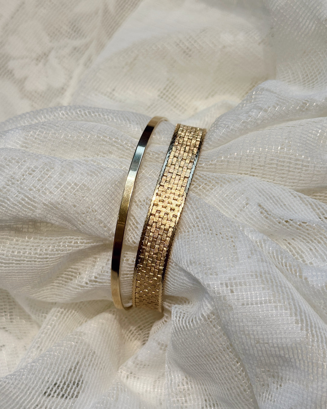 The Essential gold bangle beside a wider textured gold bracelet on gathered white fabric