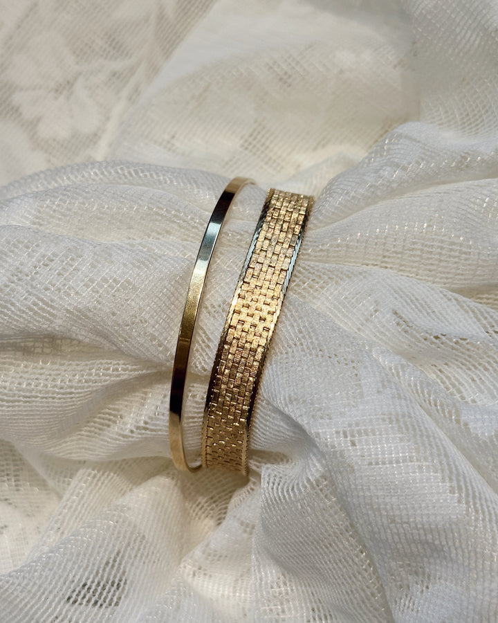 The Essential gold bangle beside a wider textured gold bracelet on gathered white fabric