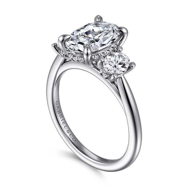 Angled view of a white gold oval three-stone engagement ring showing round side diamonds, a hidden halo and polished band.
