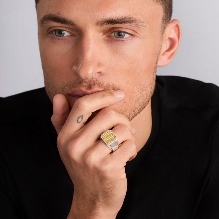 A close-up of a man with dark hair and a black shirt is holding his hand up to his chin, showcasing the ring on his wedding finger. The ring features a square gold bead motif in the middle and a thick silver band.