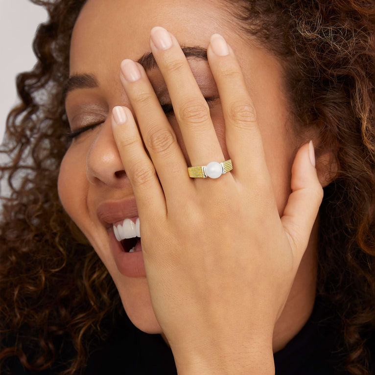 A close-up of a woman holding her hand over her face, showcasing the yellow gold ring on her middle finger. The ring features a two-tone caviar pearl ring with 18K gold beading on the band and a sterling silver form. The pearl measures 8mm in diameter.