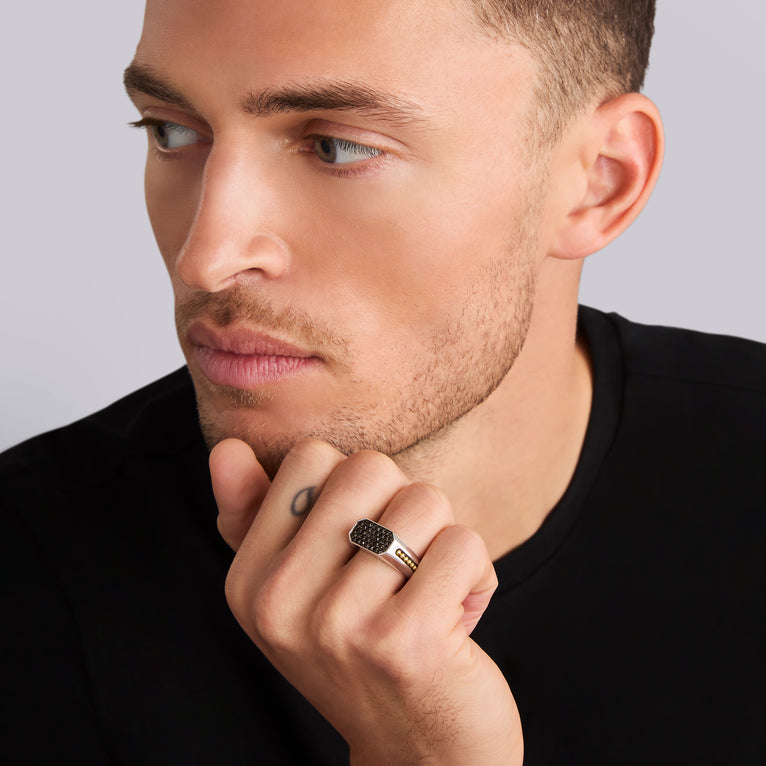 A close-up of a man holding his hand on his chin, showcasing a ring on his ring finger. The ring features an octagon-shaped center piece with Black diamonds accented by sterling silver and 18K gold Caviar beading.