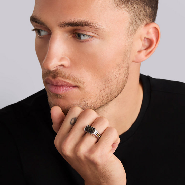 A close-up of a man holding his hand on his chin, showcasing a ring on his ring finger. The ring features an octagon-shaped center piece with Black diamonds accented by sterling silver and 18K gold Caviar beading.