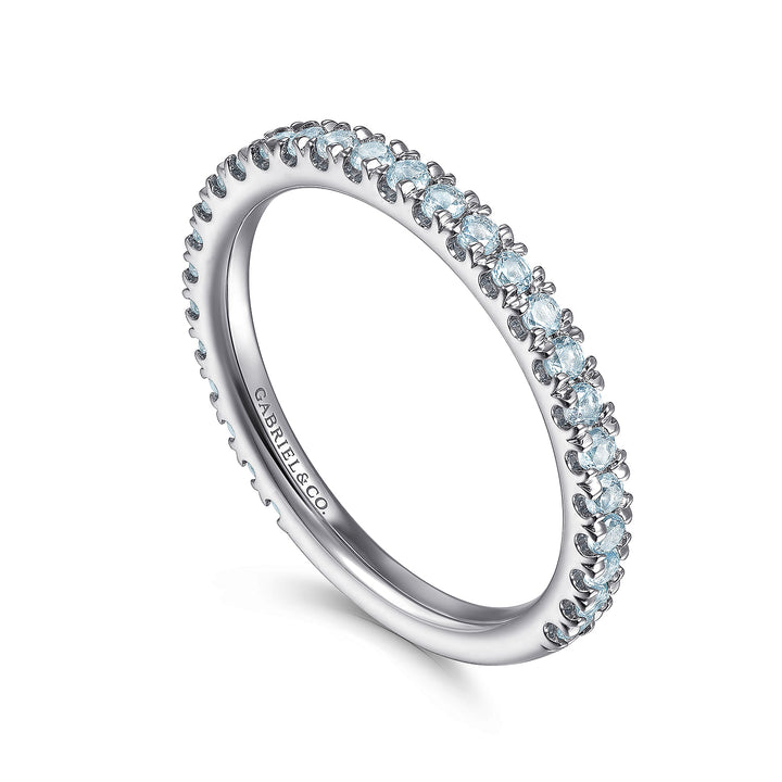 A white gold ring rests in the middle of a white background. The ring features a 3/4 eternity style with round-shaped light blue diamonds set on a band engraved with Gabriel & Co. inside the ring on the left side.