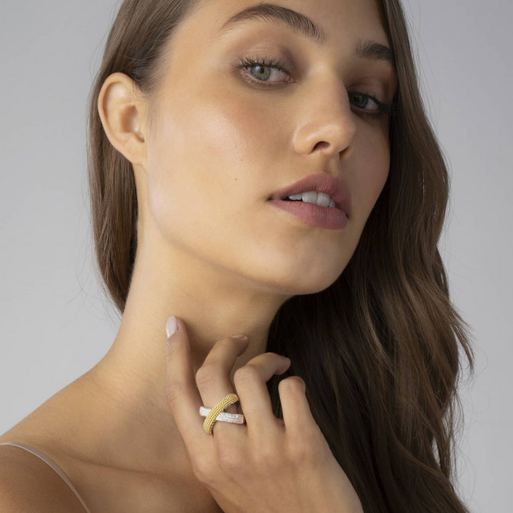 A woman with brown hair is holding her hand up to her neck, showcasing the ring on her middle finger. The ring features an x motif; one row is gold caviar beading, and the other has three rows of round-shaped diamonds.